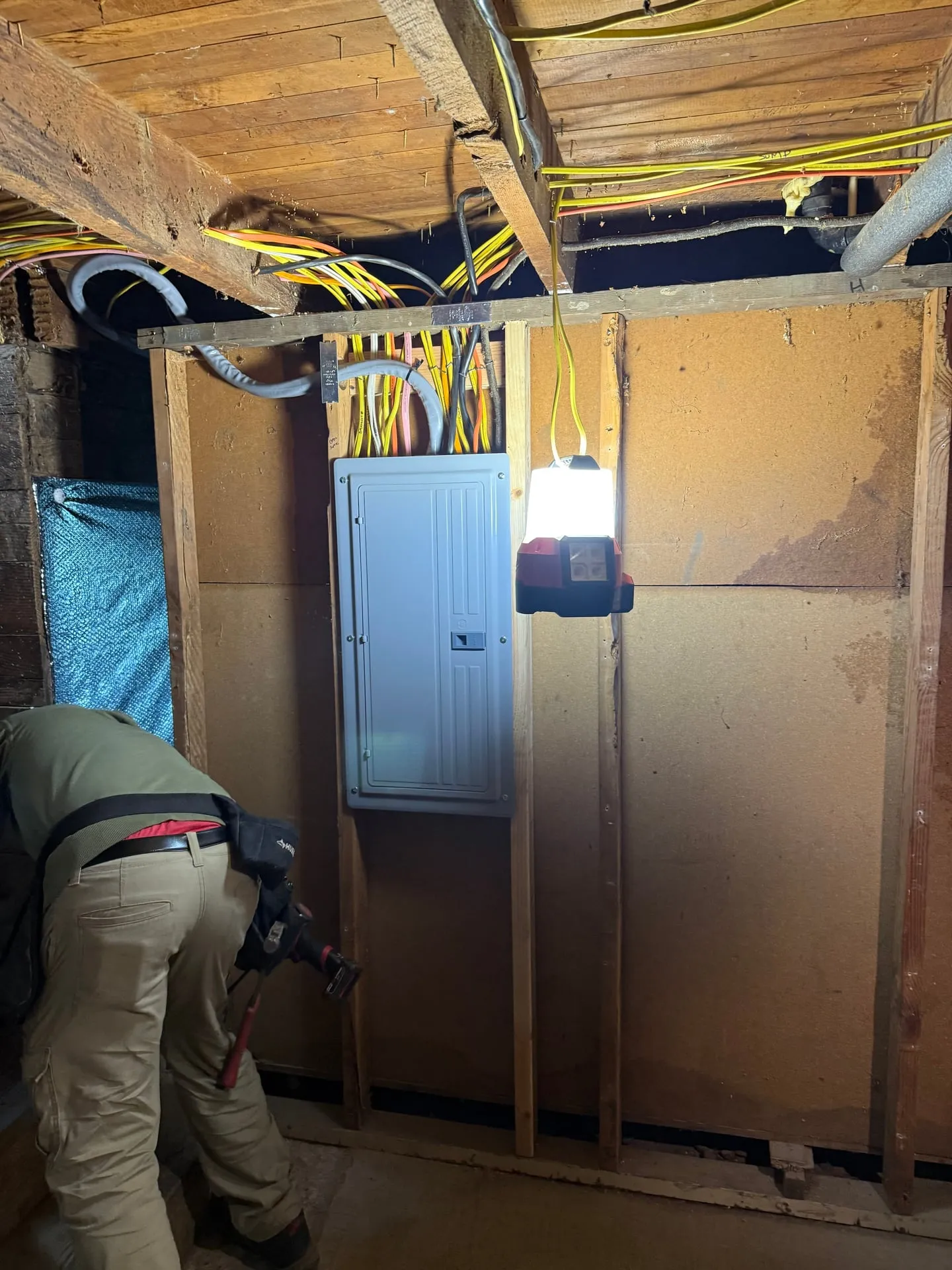 New 200-amp electrical panel mounted on a basement wall in a Ridgefield WA home with new yellow and orange NM cable dropping in from the floor joists, an electrician working at the base of the panel under a work light