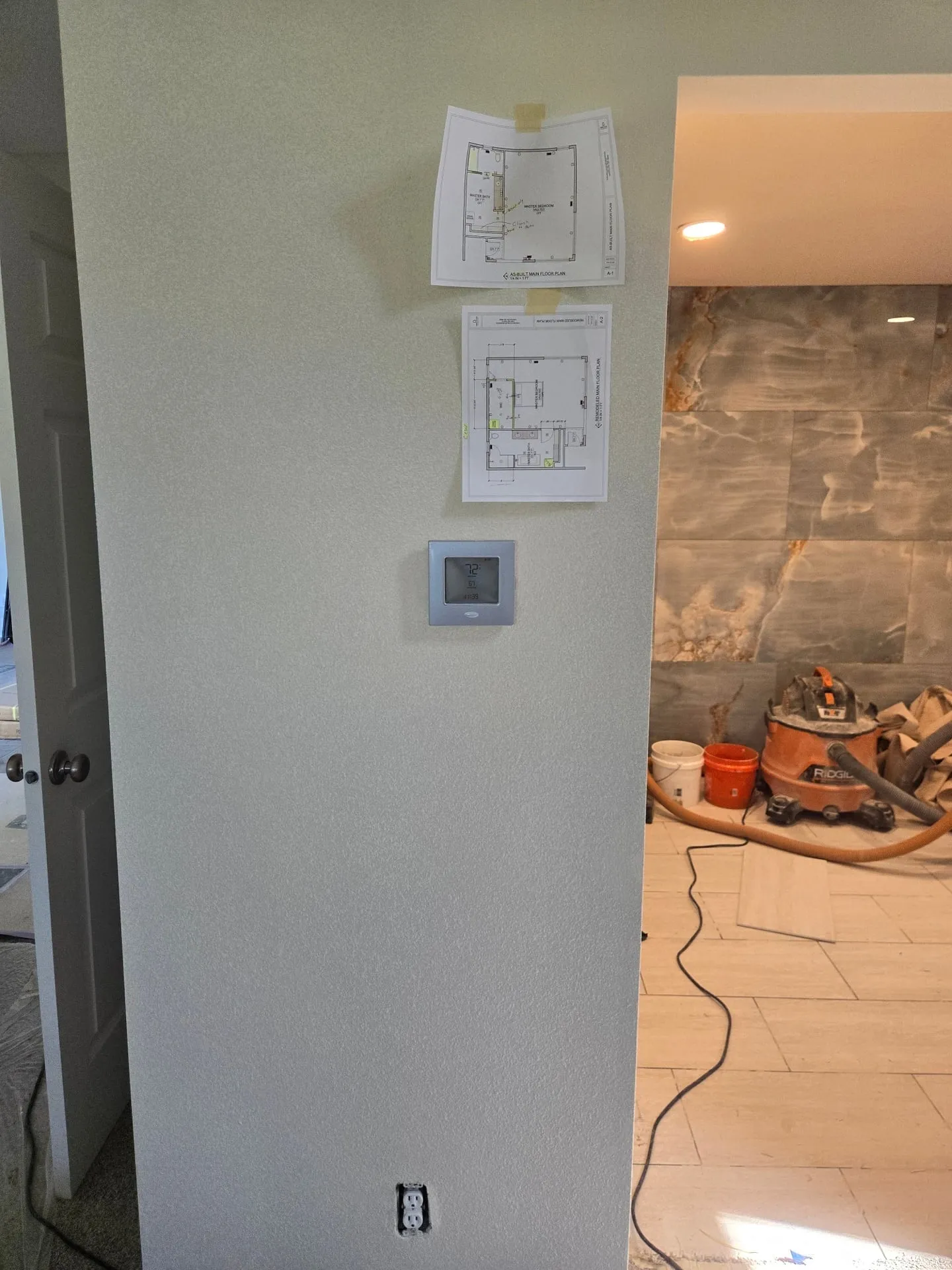 Relocated thermostat mounted on a freshly textured hallway wall next to taped-up remodel floor plans, with the tiled master shower visible through the doorway in the Orchards Vancouver WA remodel