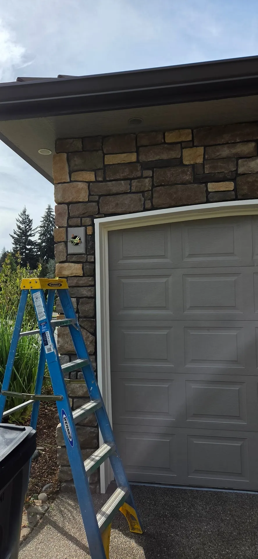 Covered electrical box on a stone veneer pilaster beside a garage door with a ladder in front, before a wall lantern was hung, at an east Vancouver WA home