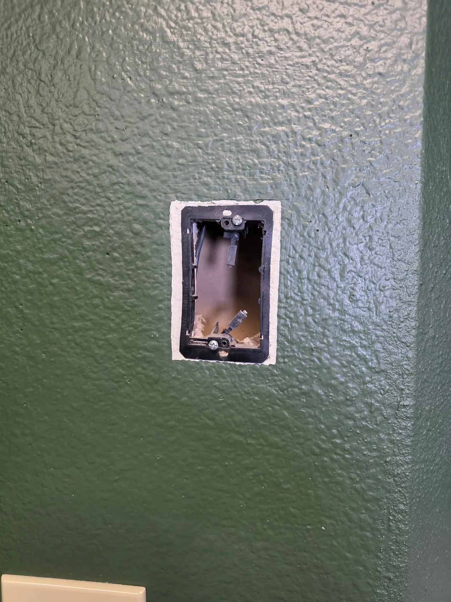 Black plastic low-voltage mounting bracket in a green wall in a Vancouver WA home, marked for low voltage use only, before a recessed outlet went in