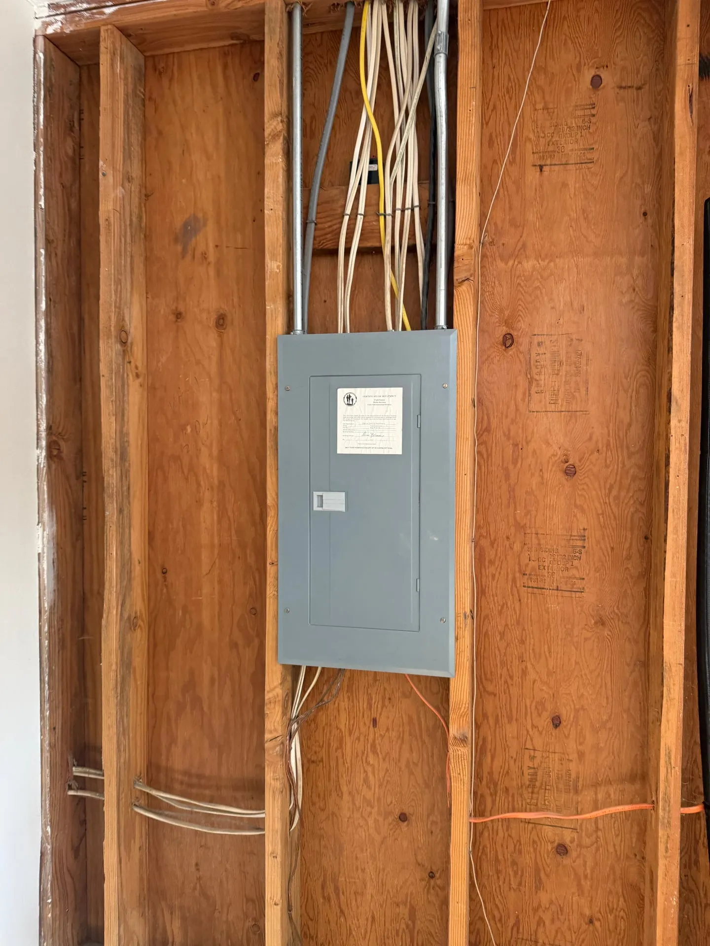 Original electrical panel in the open stud wall of a garage in Vancouver WA before replacement, with the original certificate of occupancy sticker still on the door and cables entering from above