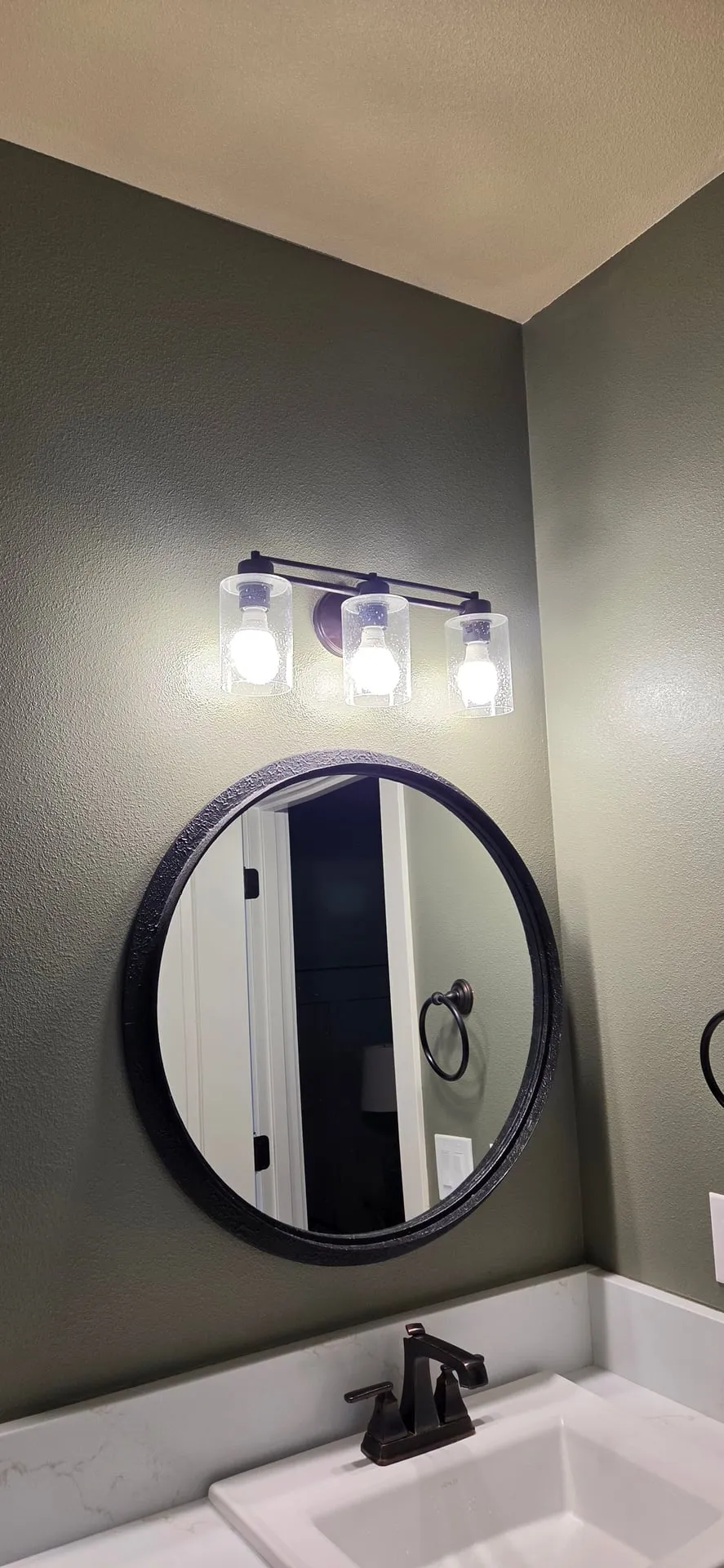 Original three-light vanity fixture with seeded glass shades and bare bulbs over a round black mirror in a Vancouver WA bathroom before replacement