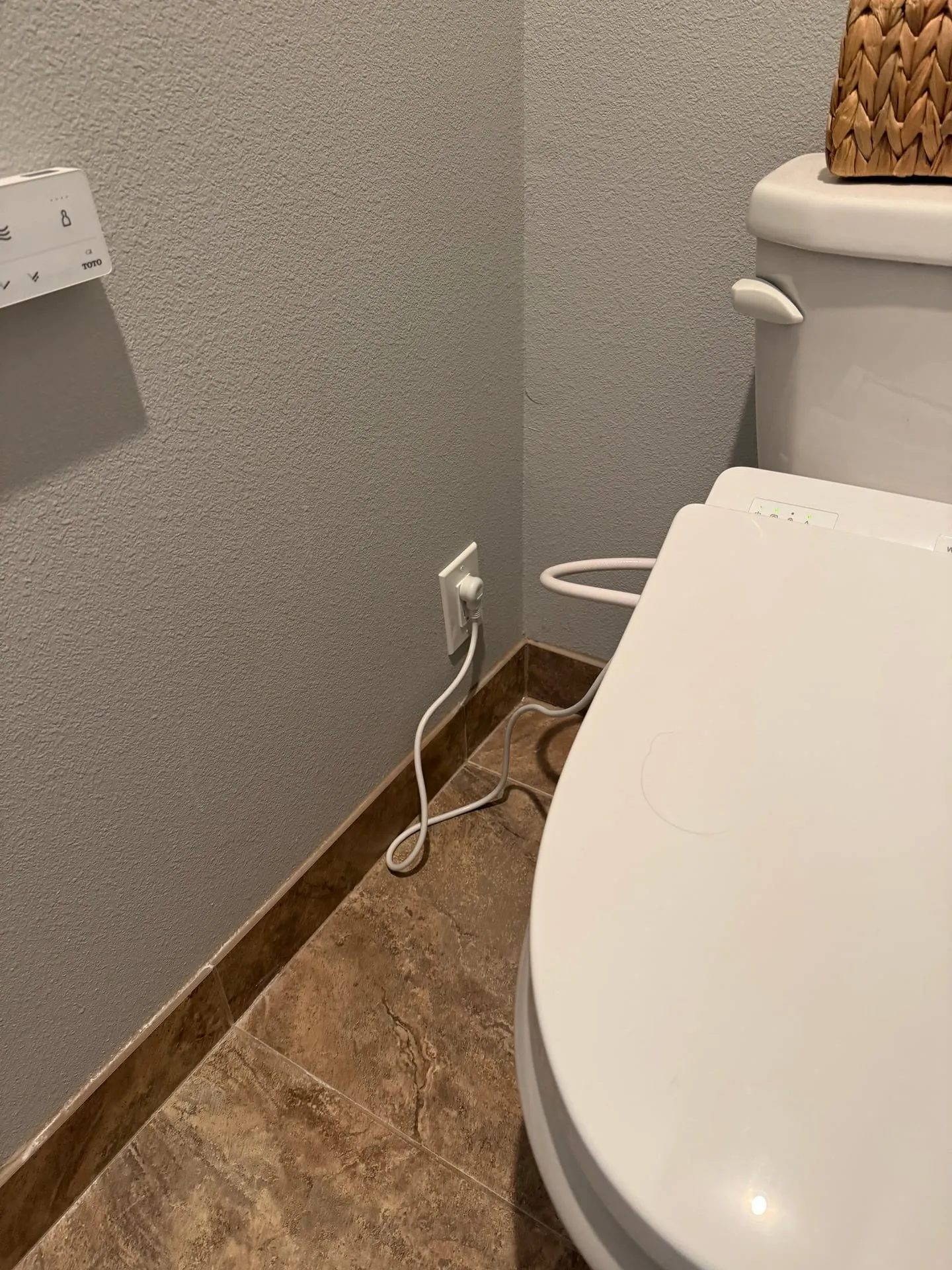 New receptacle installed low on the wall behind a toilet in a Ridgefield WA bathroom, with the bidet seat cord plugged in and routed along the baseboard
