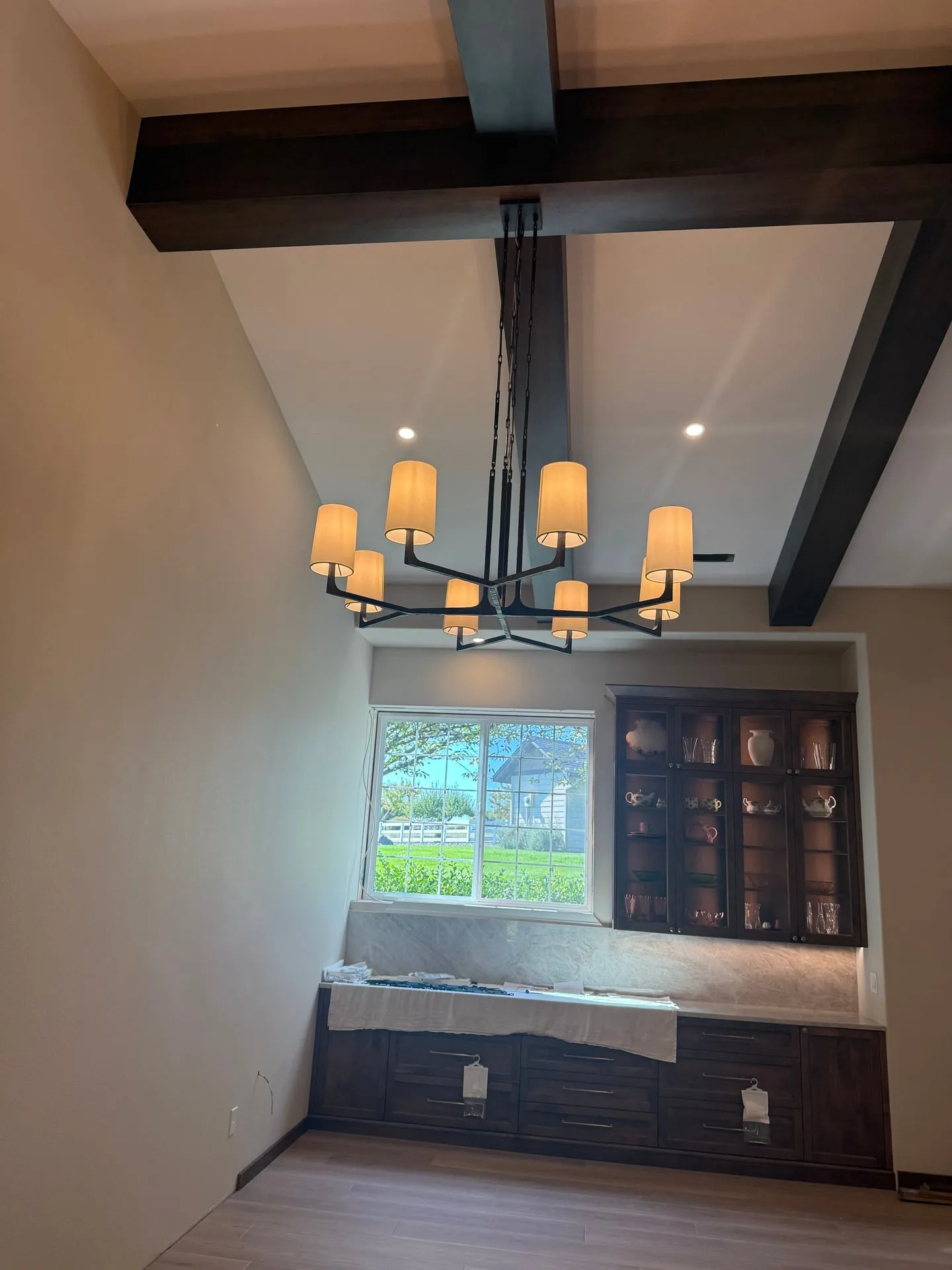 Lit chandelier with warm white shades hanging between dark beams over a built-in window seat and glass-front cabinets in a Ridgefield WA home