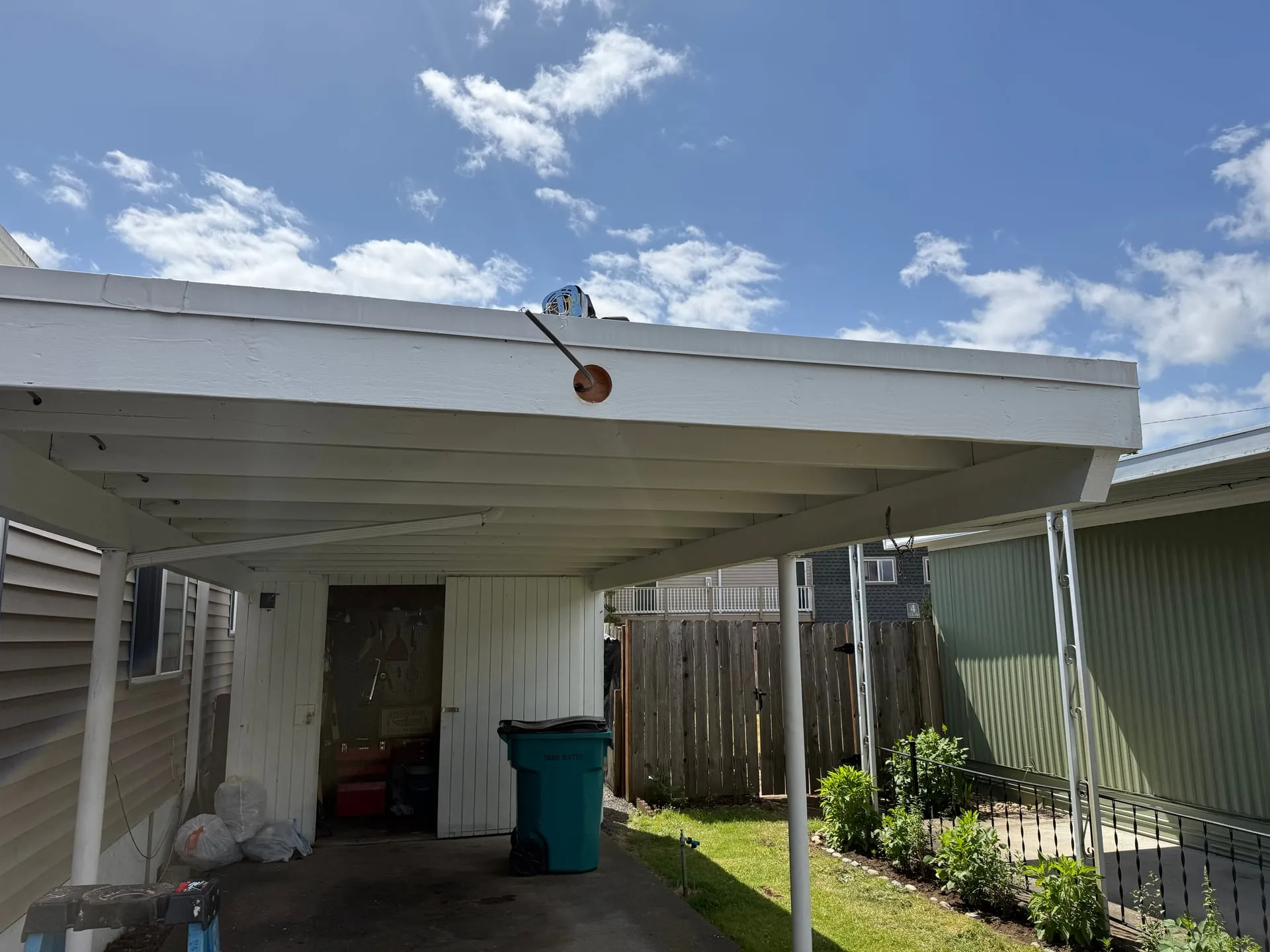 Exterior of a carport at a Vancouver WA manufactured-home community under a blue sky, with a fresh penetration drilled through the painted fascia beam and a junction box on the carport roof for a new motion-sensor light
