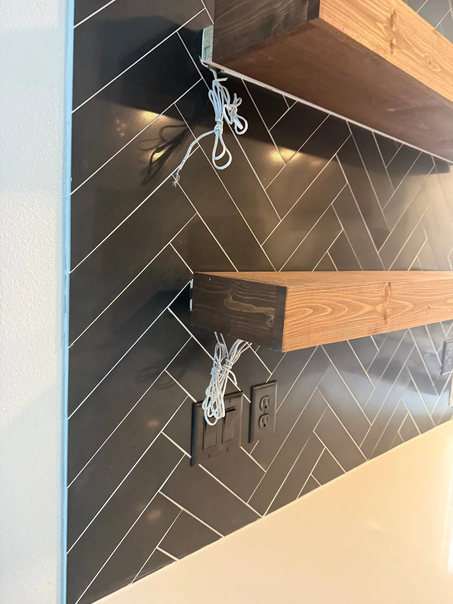 Walnut floating shelves on a black herringbone tile backsplash in a Washougal WA kitchen with white lighting leads hanging from the shelf undersides and a black outlet on the wall below