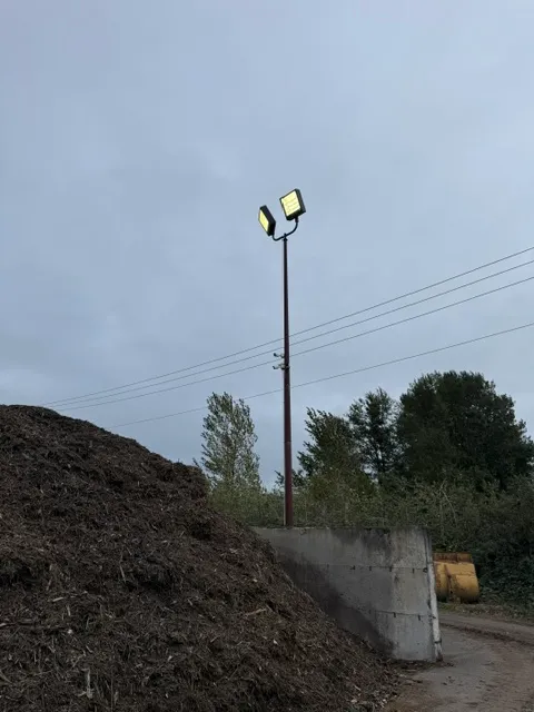 Light pole with two flood lights next to a large bark pile and concrete wall at a commercial yard in Longview WA