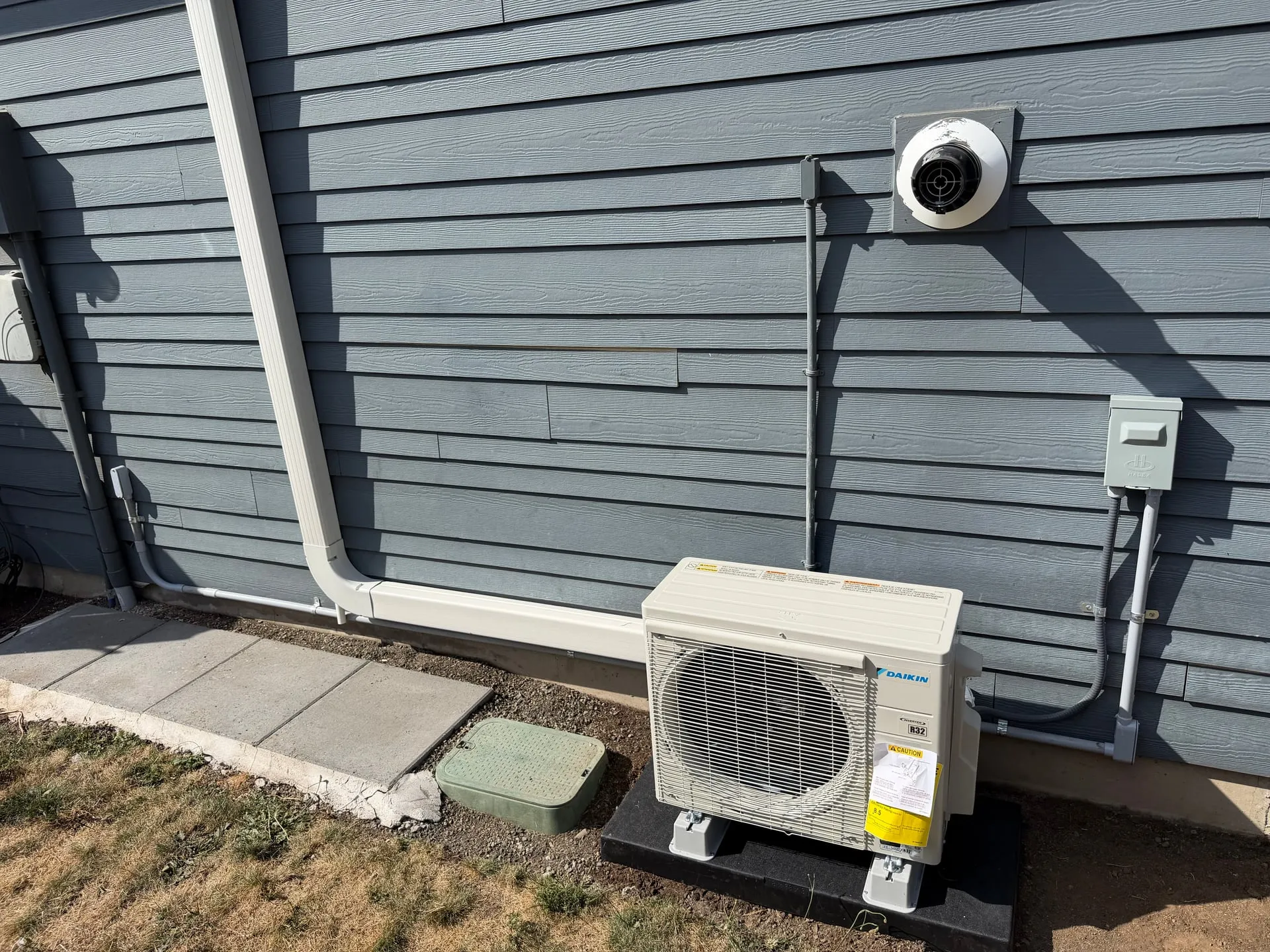 Mini split outdoor unit on a pad beside the grey lap siding of an Orchards home in Vancouver WA, with a new disconnect box and line set penetration above it