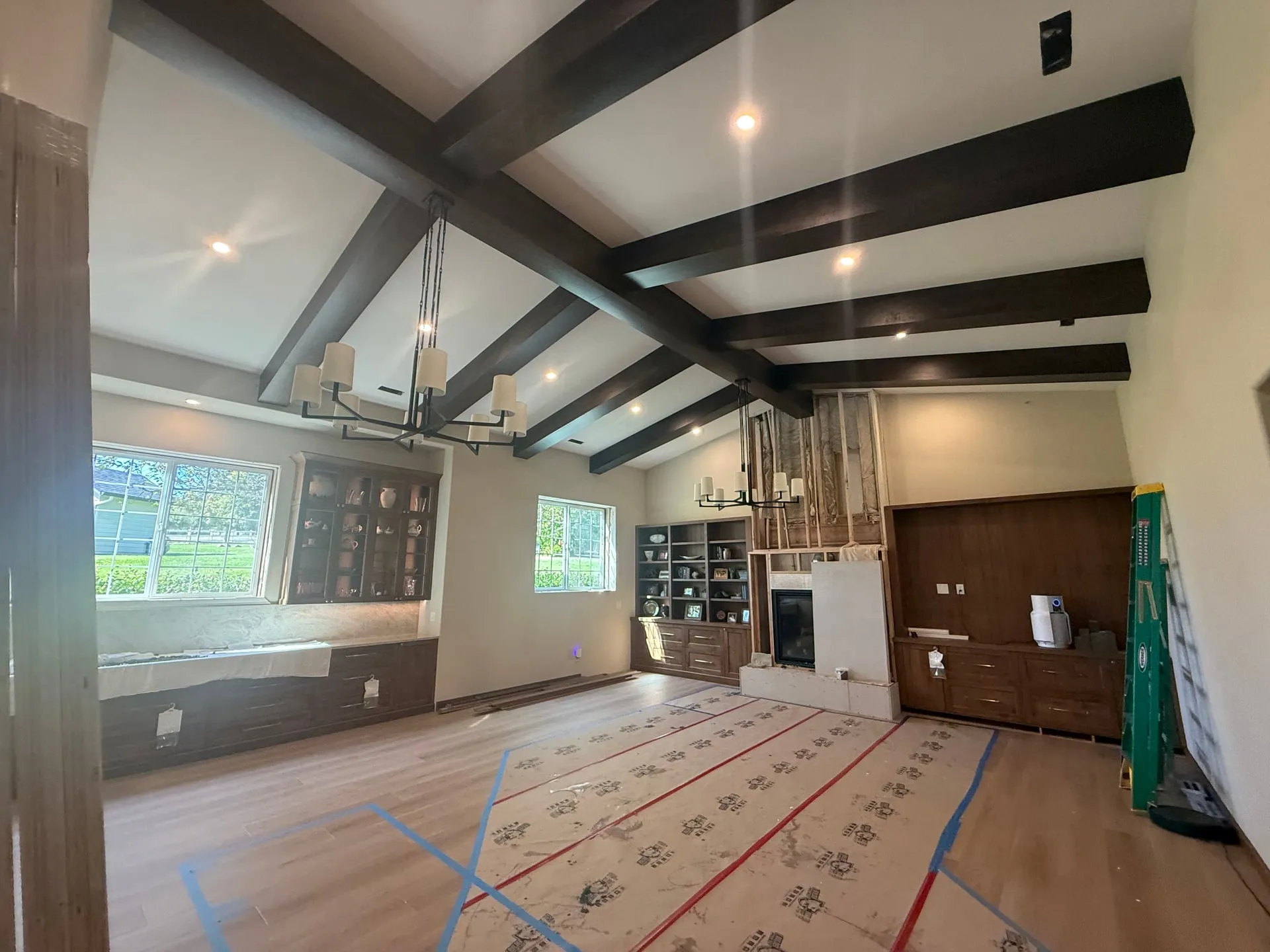 Wide view of a Ridgefield WA great room under construction showing two chandeliers hung under a vaulted ceiling with dark timber beams, built-in cabinets and a fireplace surround, with protected flooring