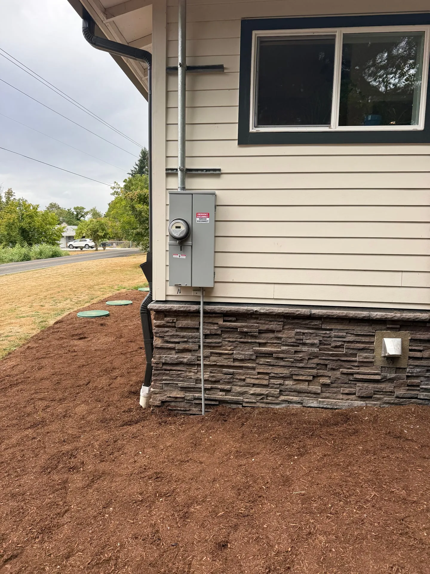 New 200-amp meter main disconnect mounted on the siding at the southwest corner of a Ridgefield WA home, with a red EMERGENCY DISCONNECT SERVICE DISCONNECT label, galvanized service mast above it and the grounding electrode conductor running down into the mulch bed