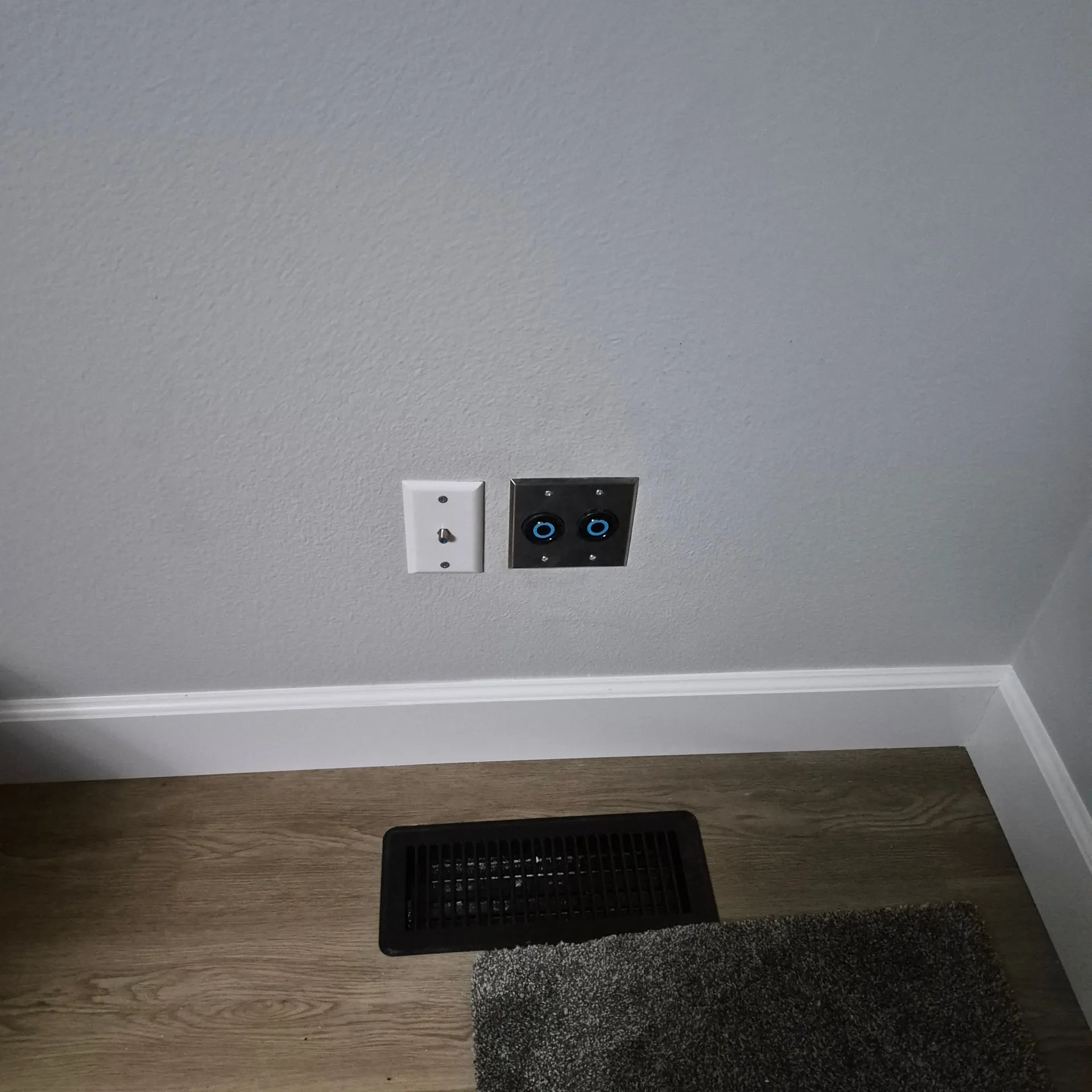 Stainless steel cover plate on a living-room wall in a Vancouver WA home holding two blue 30-amp twist-lock outlets next to a separate low-voltage jack, mounted above the baseboard near a floor vent