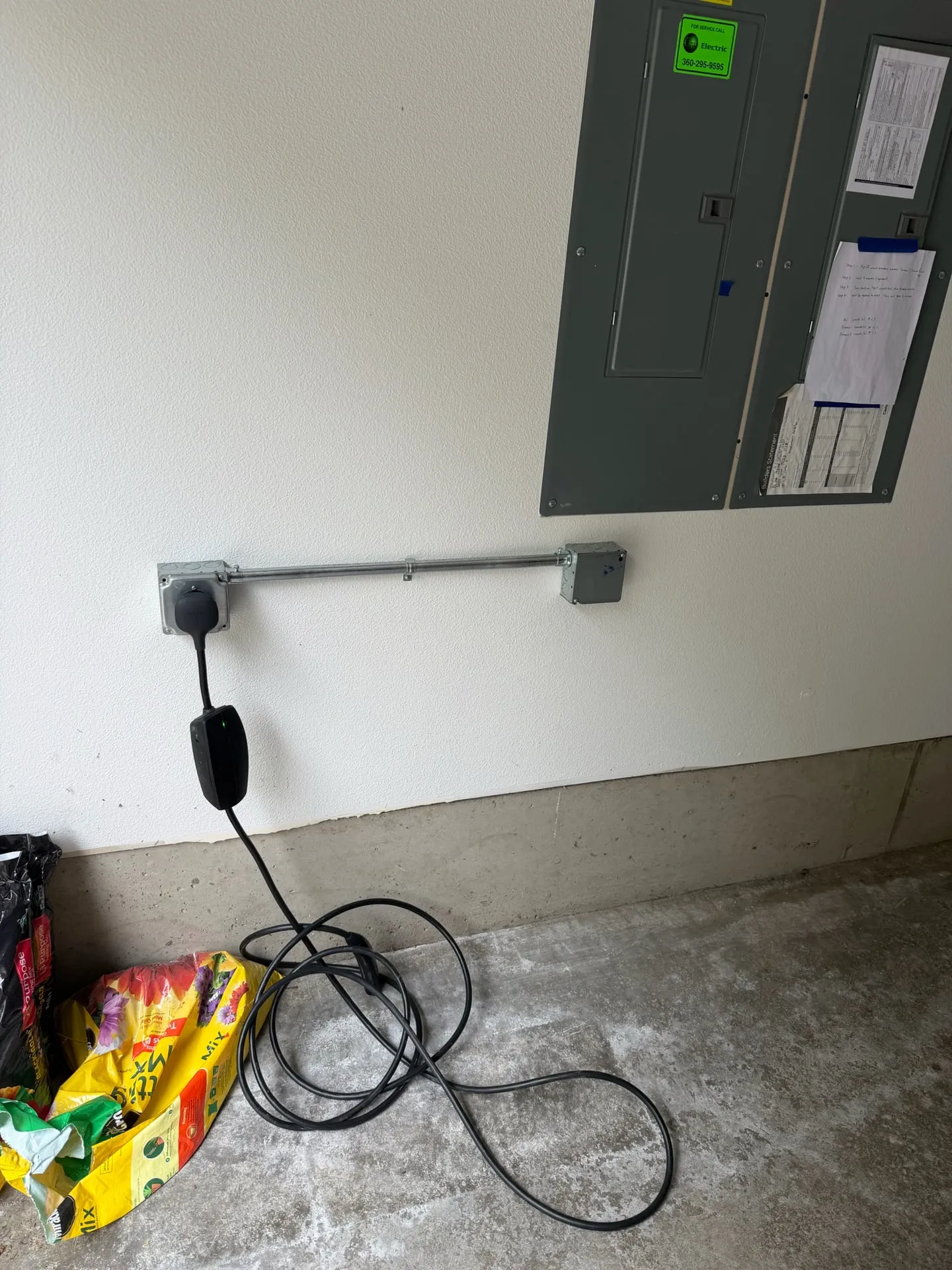 New 240-volt EV charging outlet low on a garage wall in Ridgefield WA, fed by a short horizontal run of EMT conduit from below the electrical panel, with a portable Level 2 charger plugged in and its cable coiled on the floor