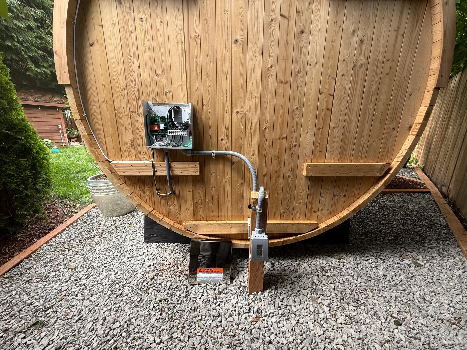 Back of a cedar barrel sauna in a Vancouver WA backyard with the Harvia power unit mounted on the exterior wall and a liquidtight conduit running down to a disconnect at the gravel base