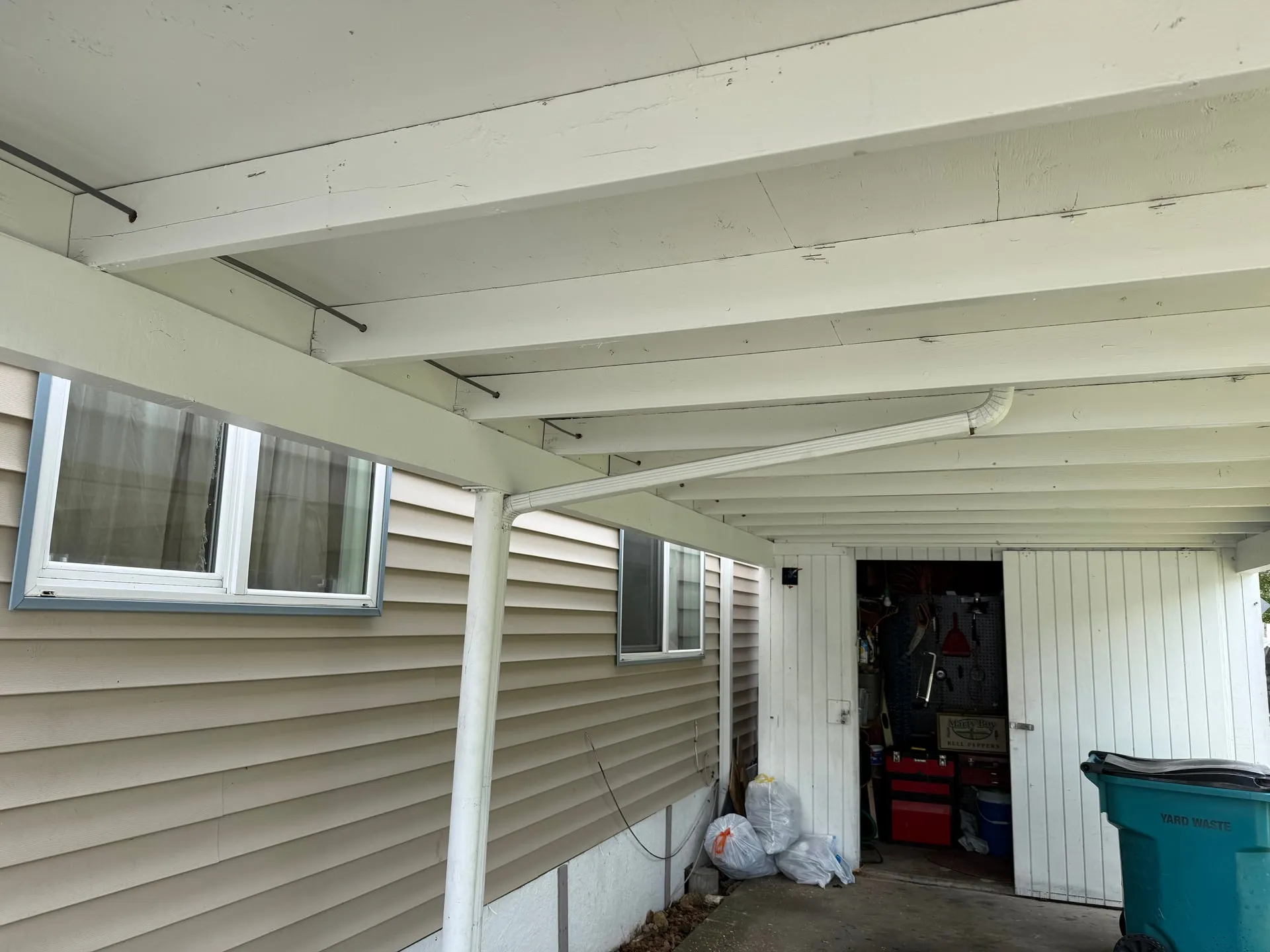 Underside of a carport roof at a Vancouver WA manufactured-home community with new surface-run EMT conduit routed along the painted beams toward a motion-sensor light location, a workshop and tool pegboard visible in the carport behind
