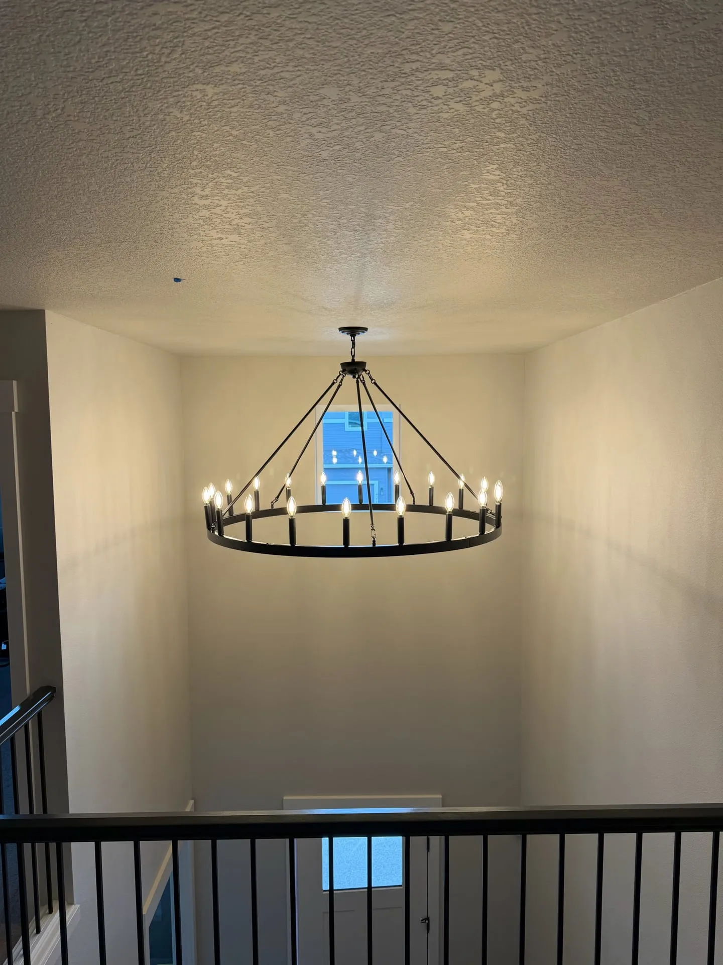 Large black wagon-wheel chandelier with candle-style lamps hanging over the open stairwell landing of a Salmon Creek home in Vancouver WA, seen from across the landing with the vaulted ceiling above