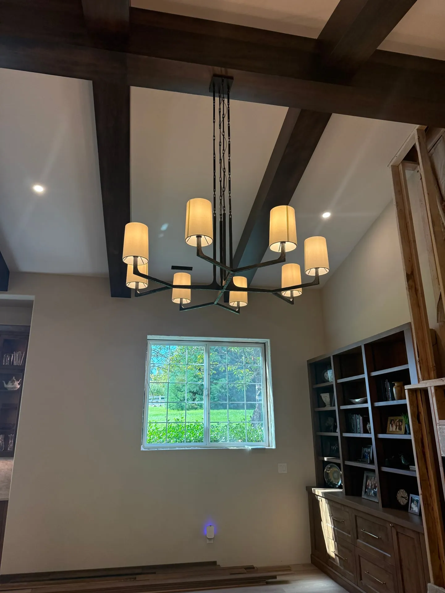 Black multi-arm chandelier with white linen shades hanging from a vaulted ceiling between dark timber beams in a Ridgefield WA great room, with recessed lights in the ceiling and built-in dark wood shelving below