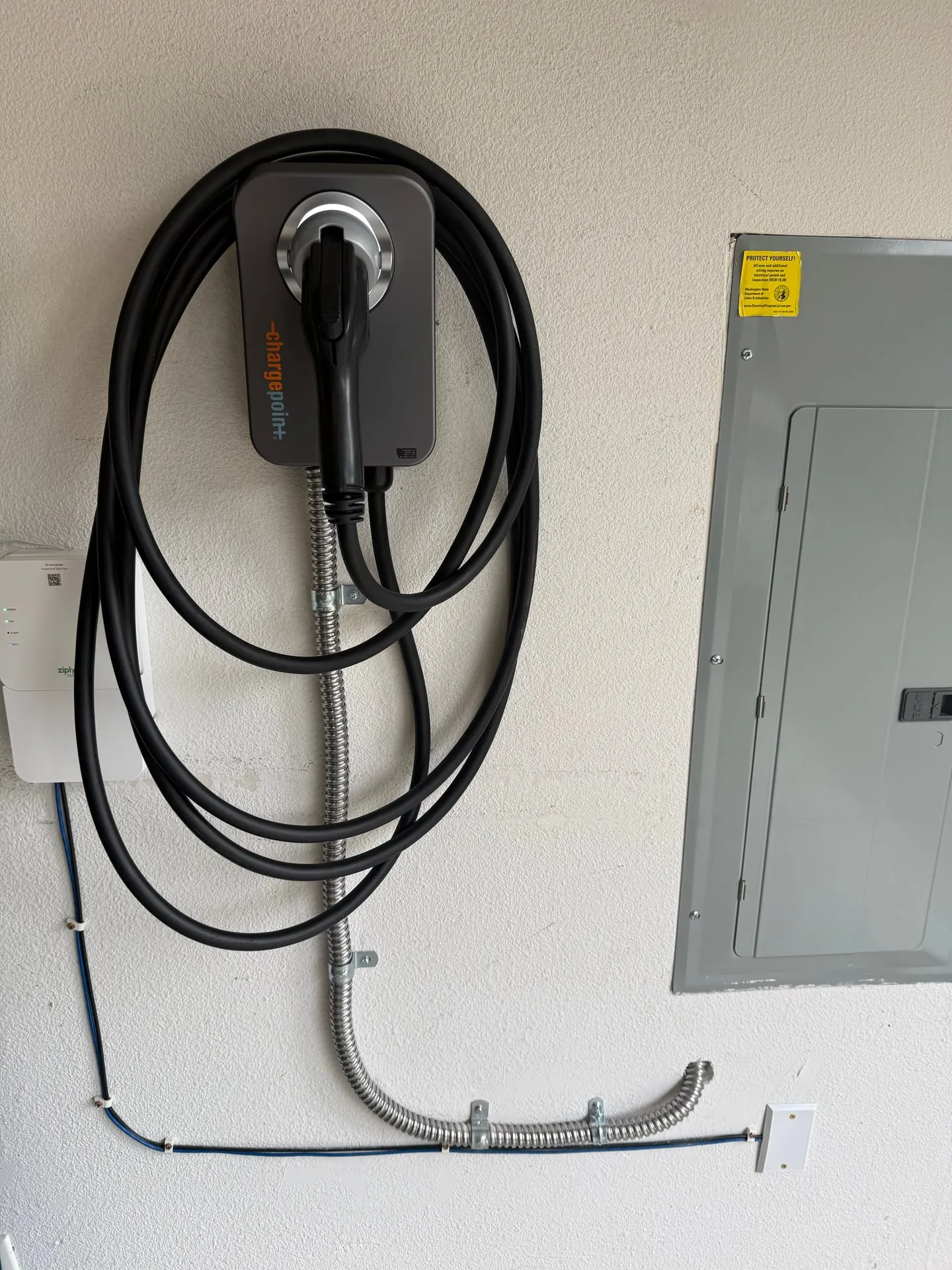 Hardwired ChargePoint EV charger mounted on the garage wall of a Camas WA home with its cable coiled on the holster, fed by a short flexible whip from the panel a few feet to the right