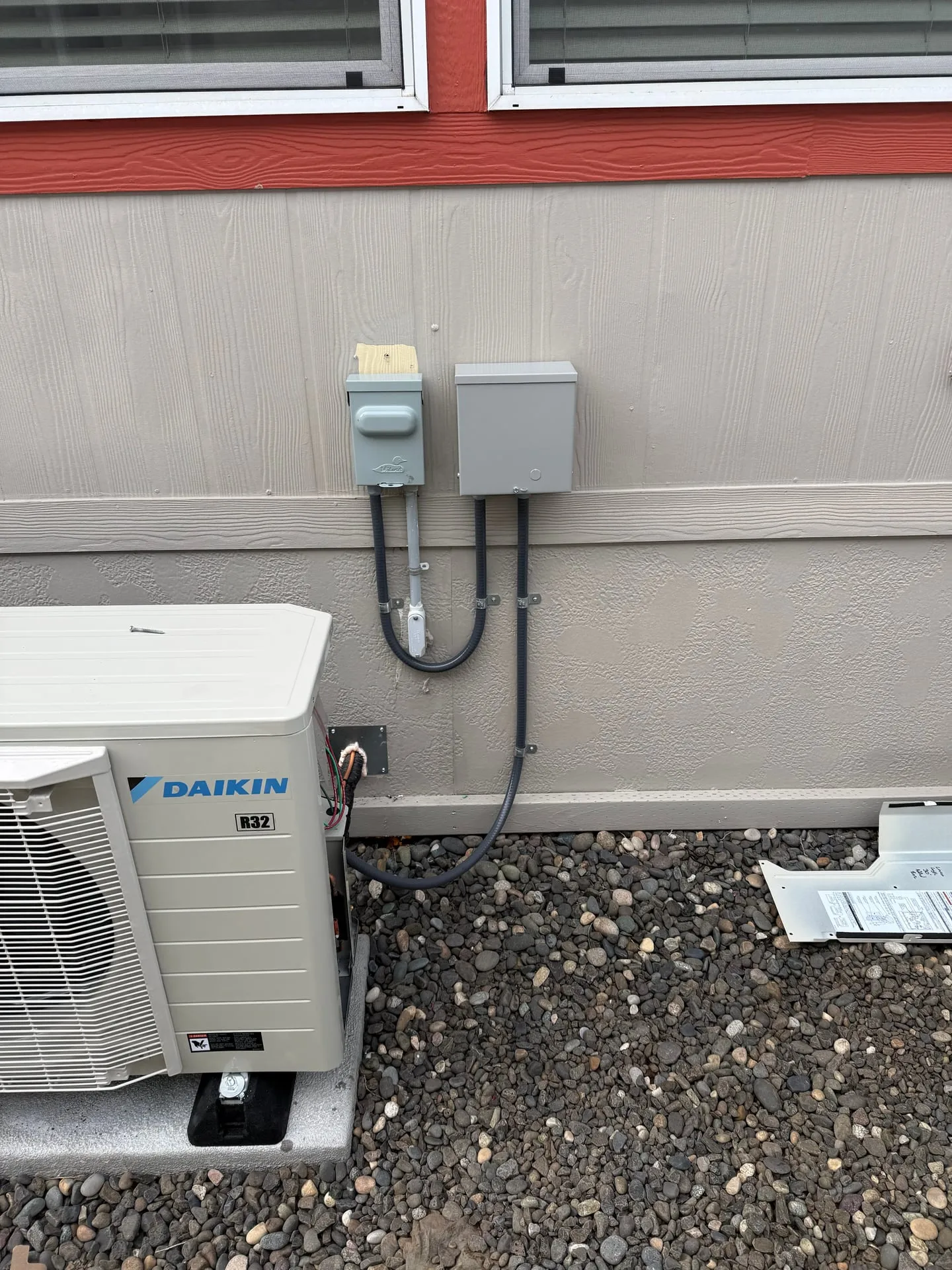 Daikin outdoor heat pump unit on a gravel pad beside a manufactured home in Orchards, Vancouver WA, with a new disconnect box mounted on the skirting wall above and whip running down to the unit