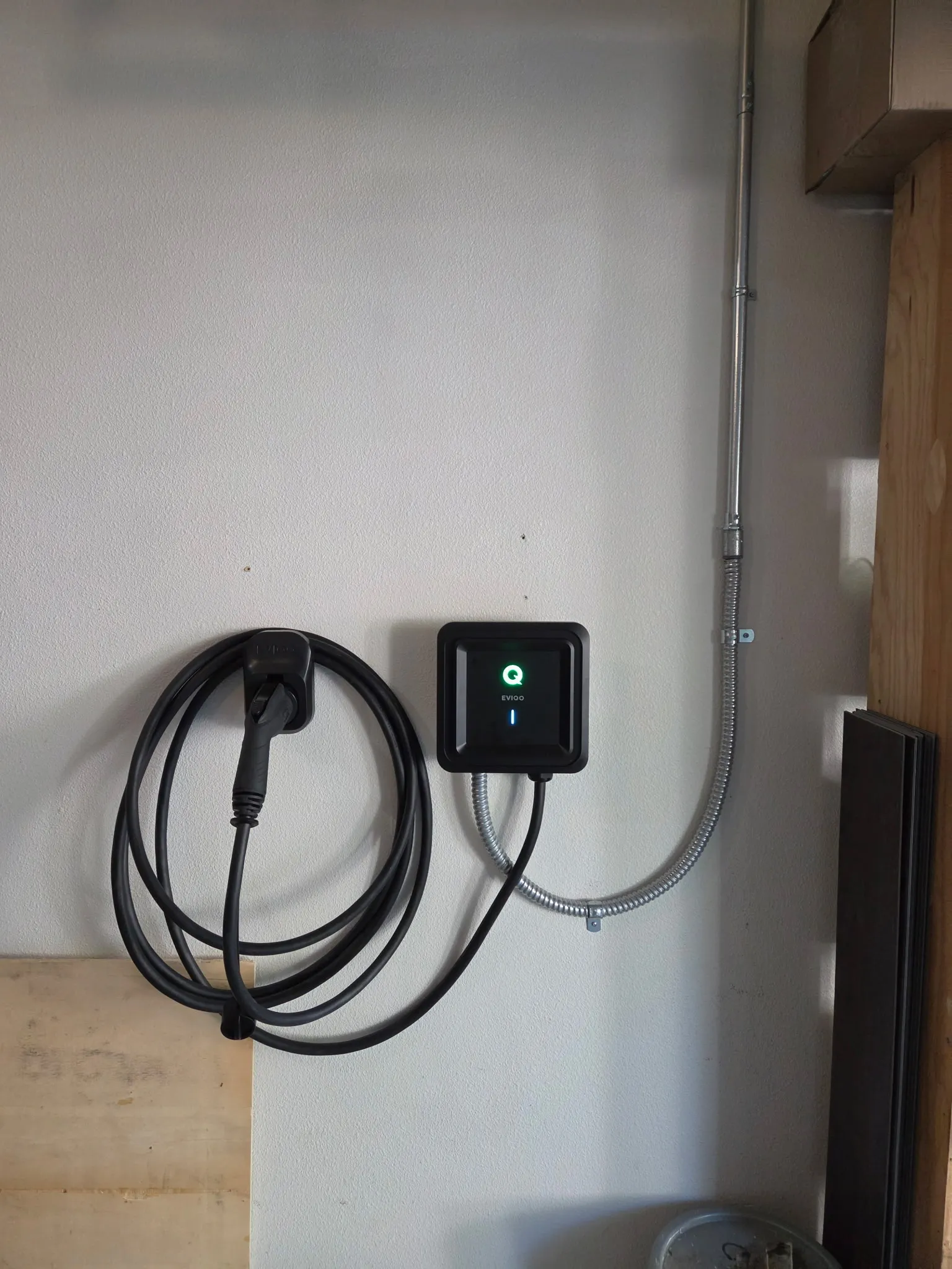 Hardwired EVIQO Level 2 EV charger mounted on a garage wall in Vancouver WA with its charging cable coiled on a holster, fed by EMT conduit down the wall and a short flexible metal whip into the bottom of the unit