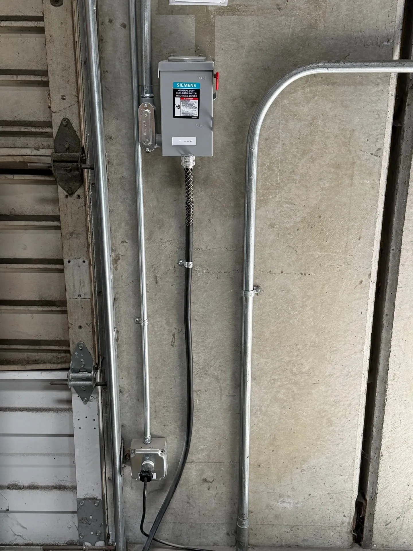 Fused safety disconnect mounted on the tilt-up concrete wall of a Vancouver WA warehouse, fed by rigid conduit running up the wall with a receptacle below for the machine