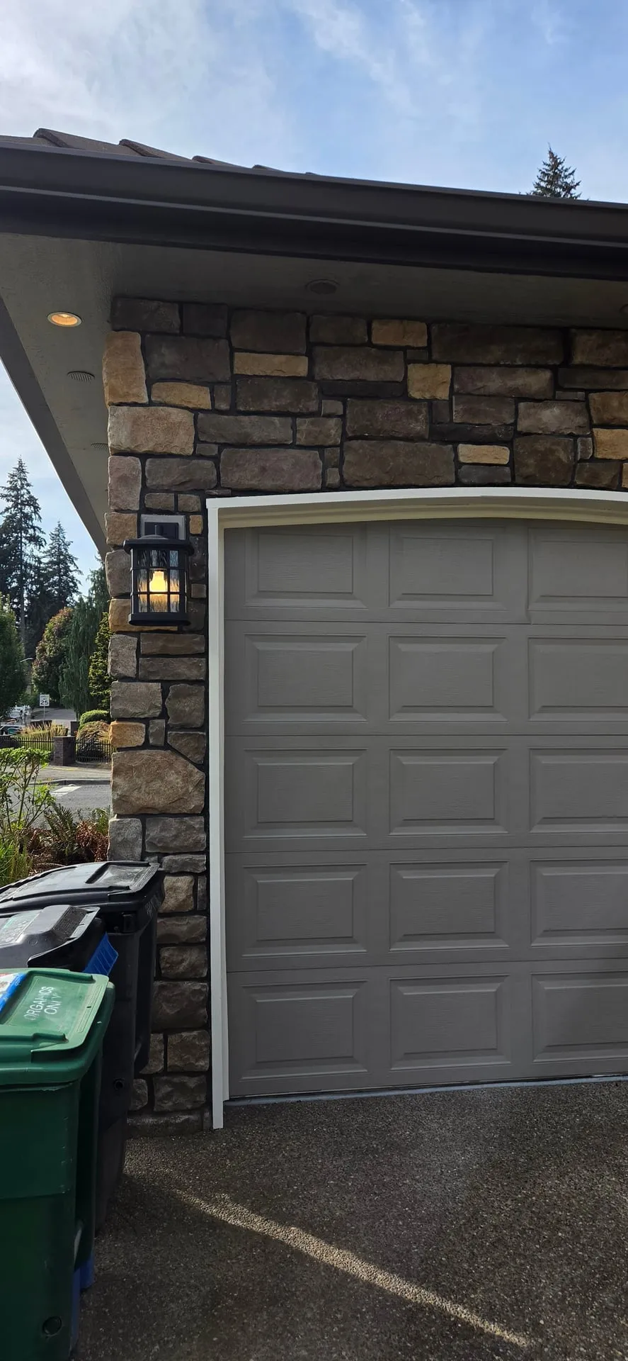 New black wall lantern with seeded glass lit on a stone veneer pilaster beside a gray garage door on an east Vancouver WA home