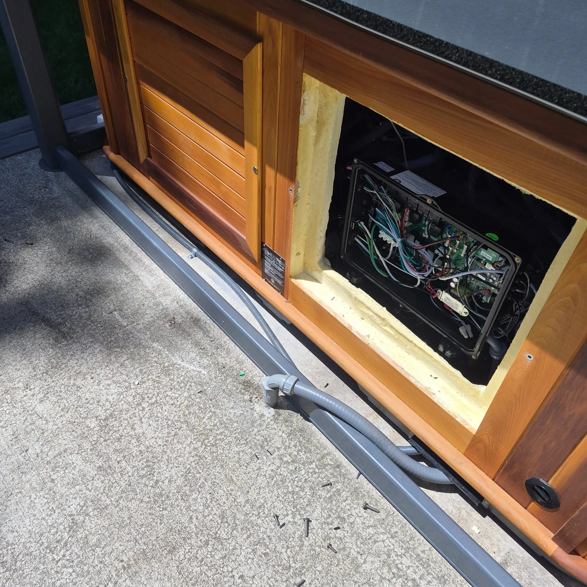 Cedar-cabinet hot tub at a Brush Prairie WA home with the equipment bay open to show the spa pack control board, and a gray liquidtight conduit whip running along the base of the cabinet to feed it