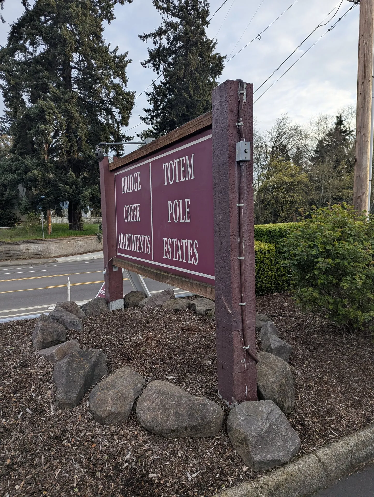Roadside monument sign reading Bridge Creek Apartments and Totem Pole Estates in Vancouver WA, with newly installed low-voltage lighting fixtures mounted on arms above the sign face