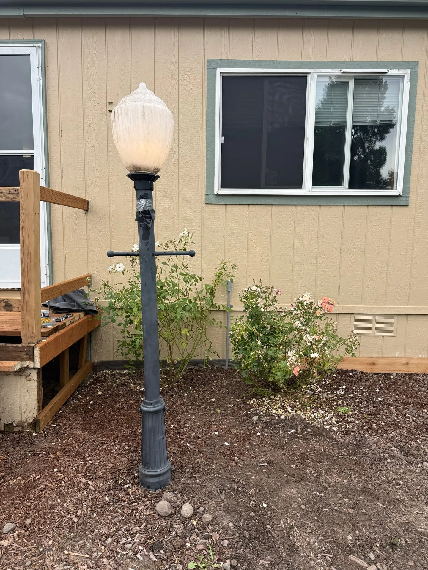 Restored black cast iron post lamp lit outside an Orchards home in Vancouver WA, with its fluted glass globe back in place, standing in a fresh planting bed beside the deck