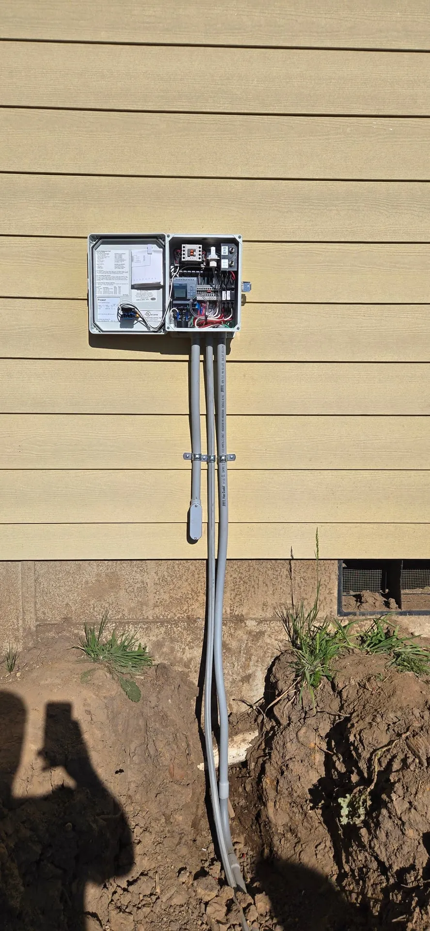 SunWater septic treatment control panel mounted on the yellow lap siding of a new-construction home in Kalama WA, with conduit running down the wall into the ground toward the tank risers