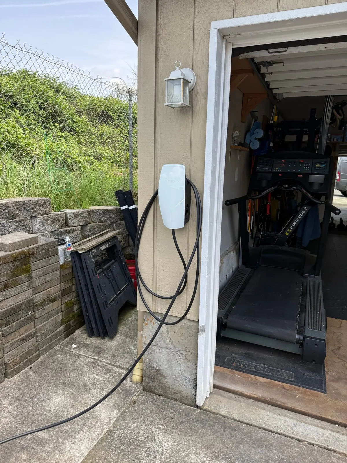 Tesla Wall Connector mounted on the exterior garage siding of a Minnehaha home in Vancouver WA, charging cable coiled and trailing down to the concrete driveway, with an outdoor sconce above and a retaining wall in the foreground