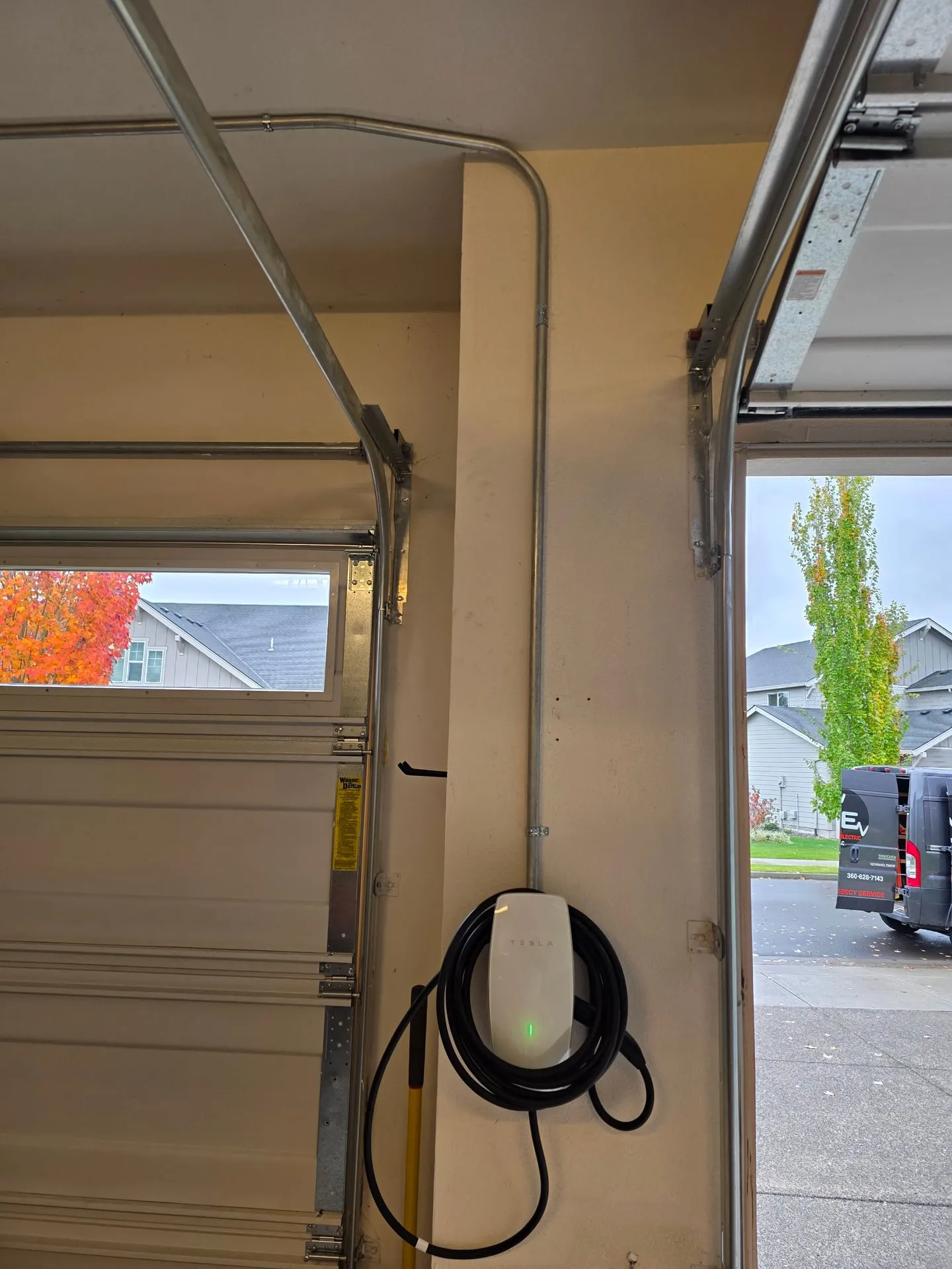 White Tesla Wall Connector with its cable coiled, mounted on the garage wall beside an open garage door in a Vancouver WA home, fed by metal conduit