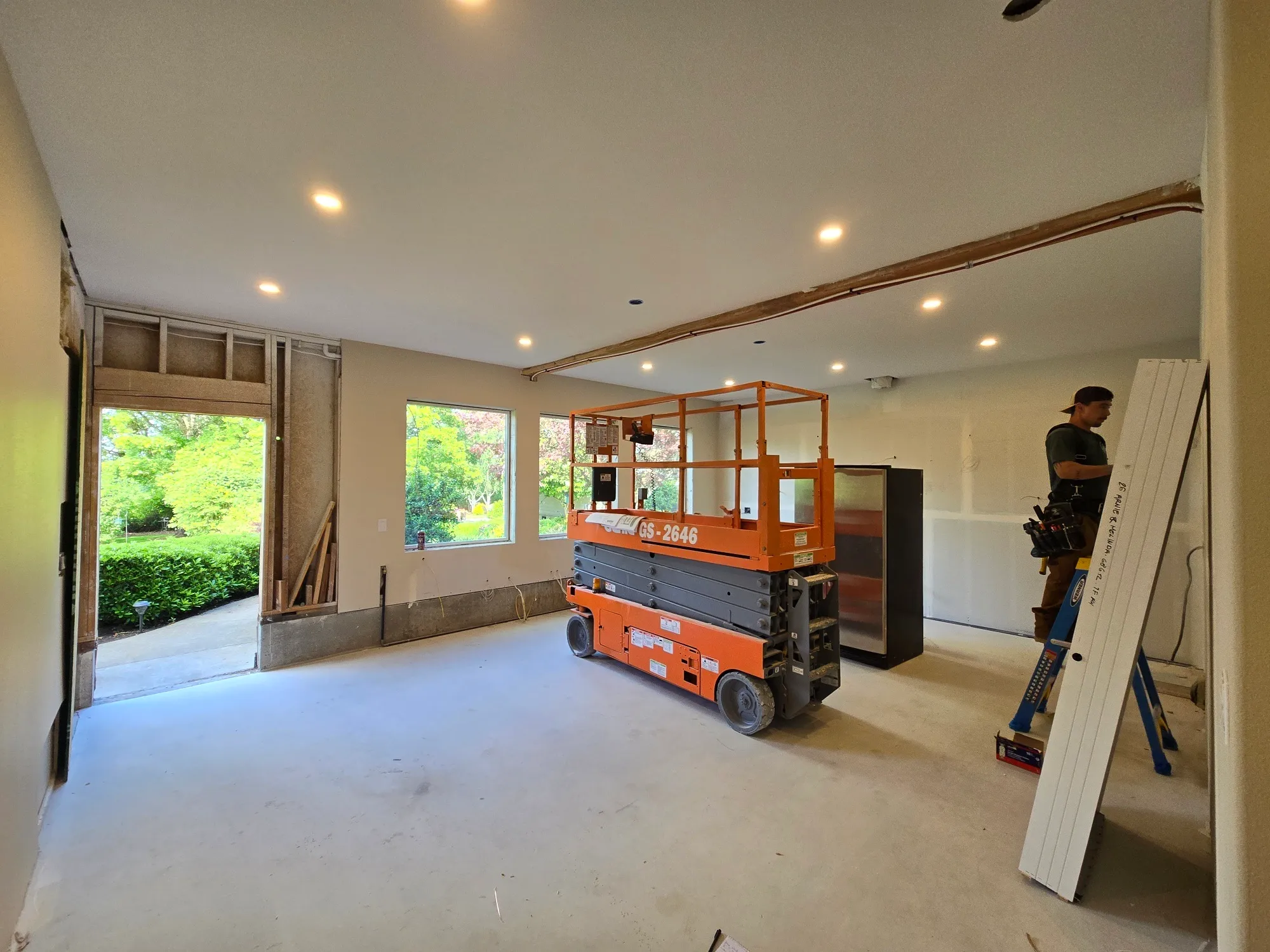 Open great room remodel in a Battle Ground WA home with new recessed can lights installed across a high vaulted ceiling, an orange scissor lift parked mid-room and an electrician in a tool belt working off a ladder near the framed wall opening