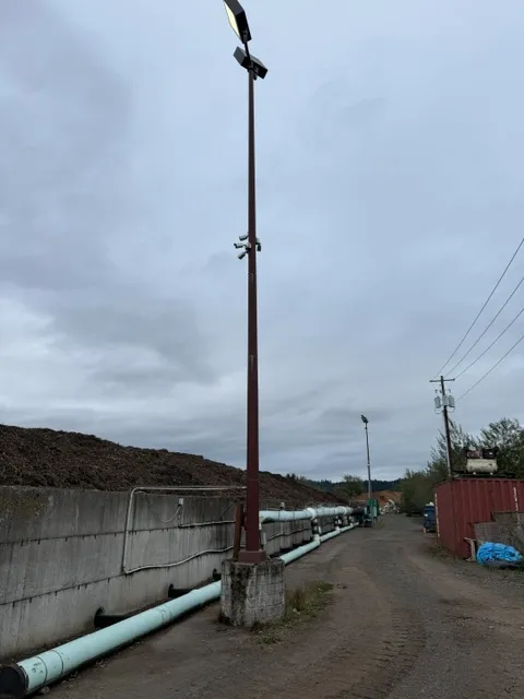 Tall light pole with flood lights along a gravel road beside a concrete block wall and piping at a commercial yard in Longview WA