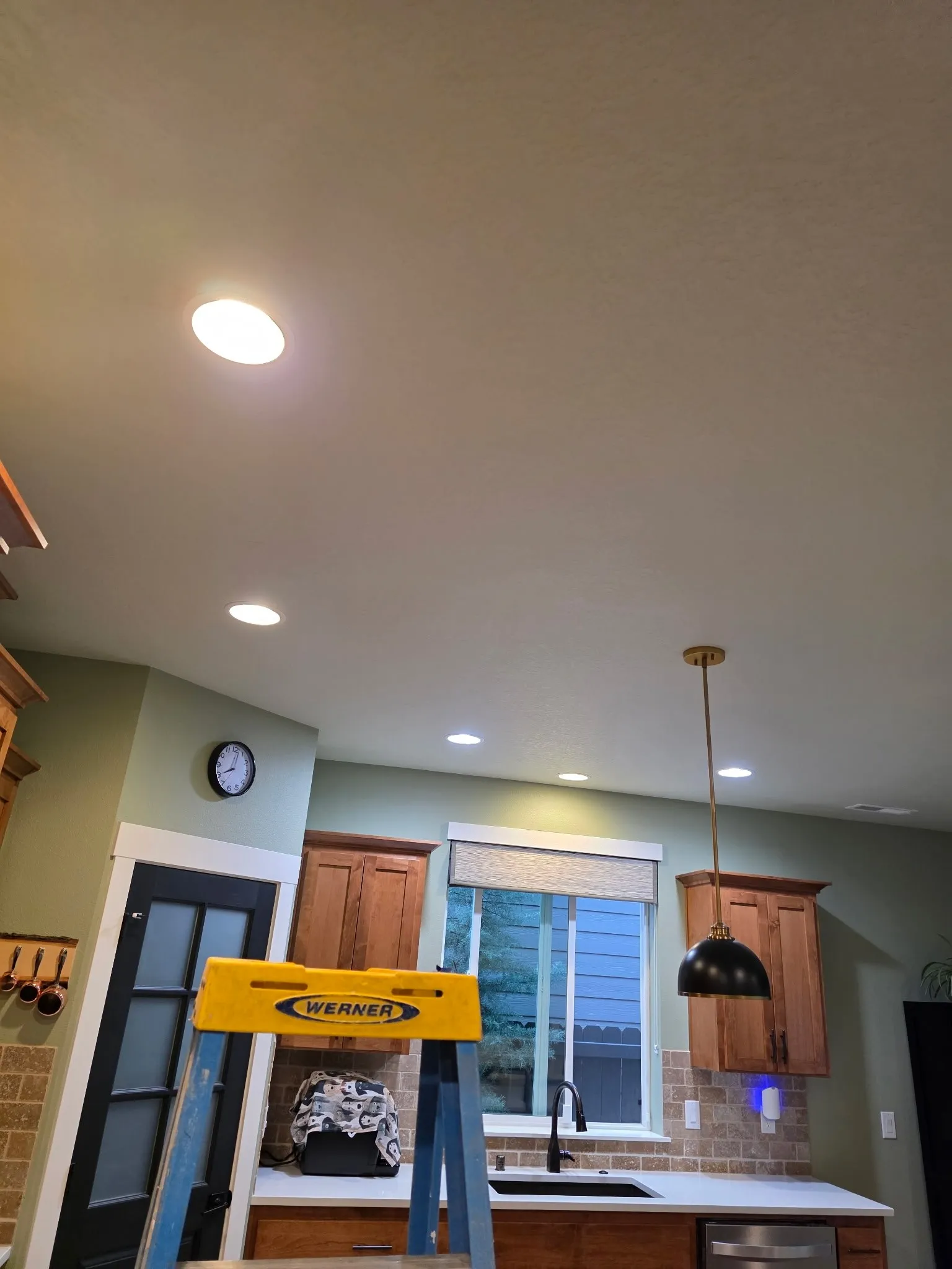Kitchen in a Vancouver WA home with LED retrofit trims in the existing 6-inch recessed cans, a black dome pendant over the sink and maple cabinets, with a ladder in the foreground