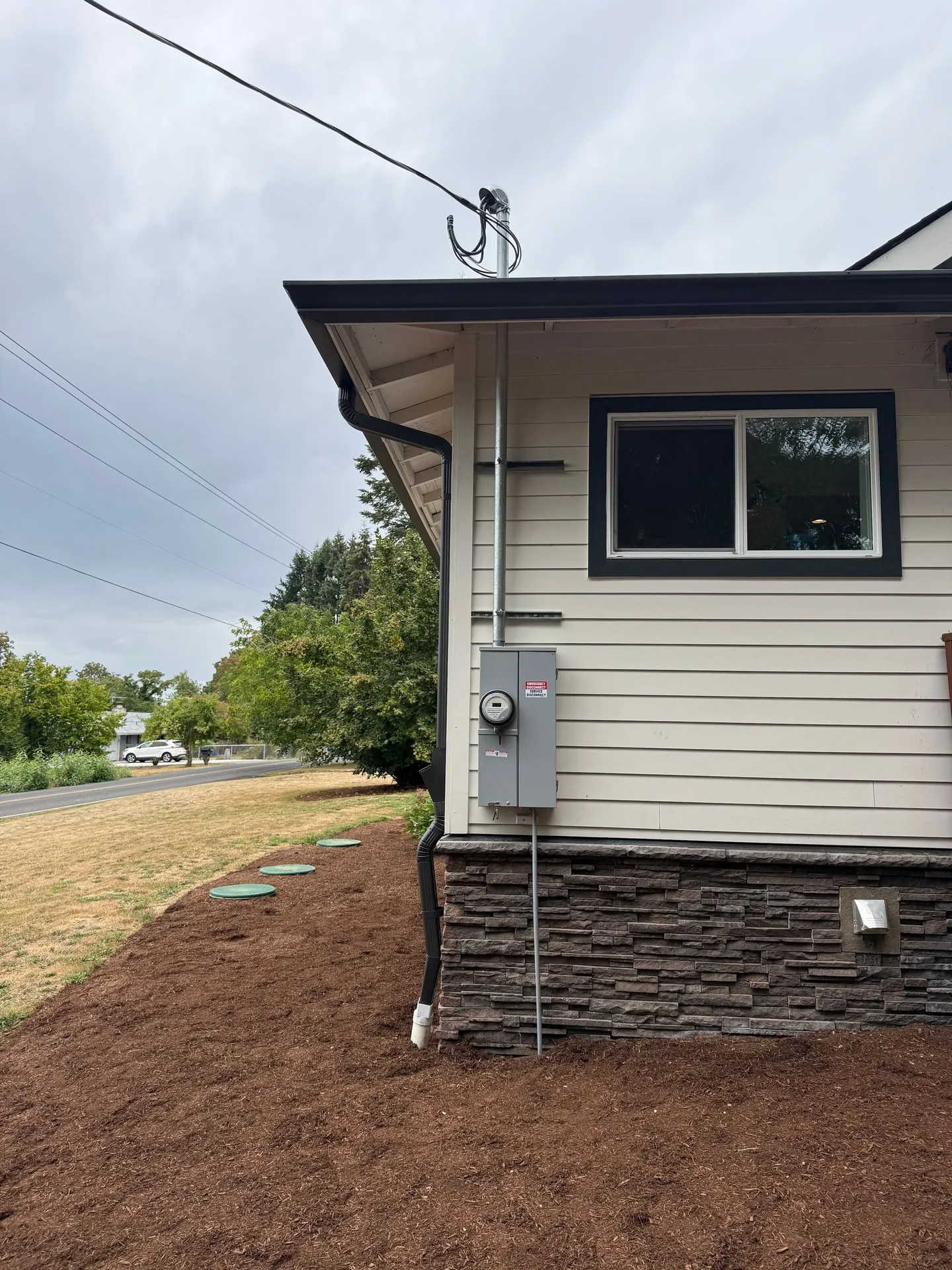Overhead service drop landing on a new galvanized mast at the southwest corner of a Ridgefield WA home, feeding the 200-amp meter main mounted on the siding above a stone veneer skirt