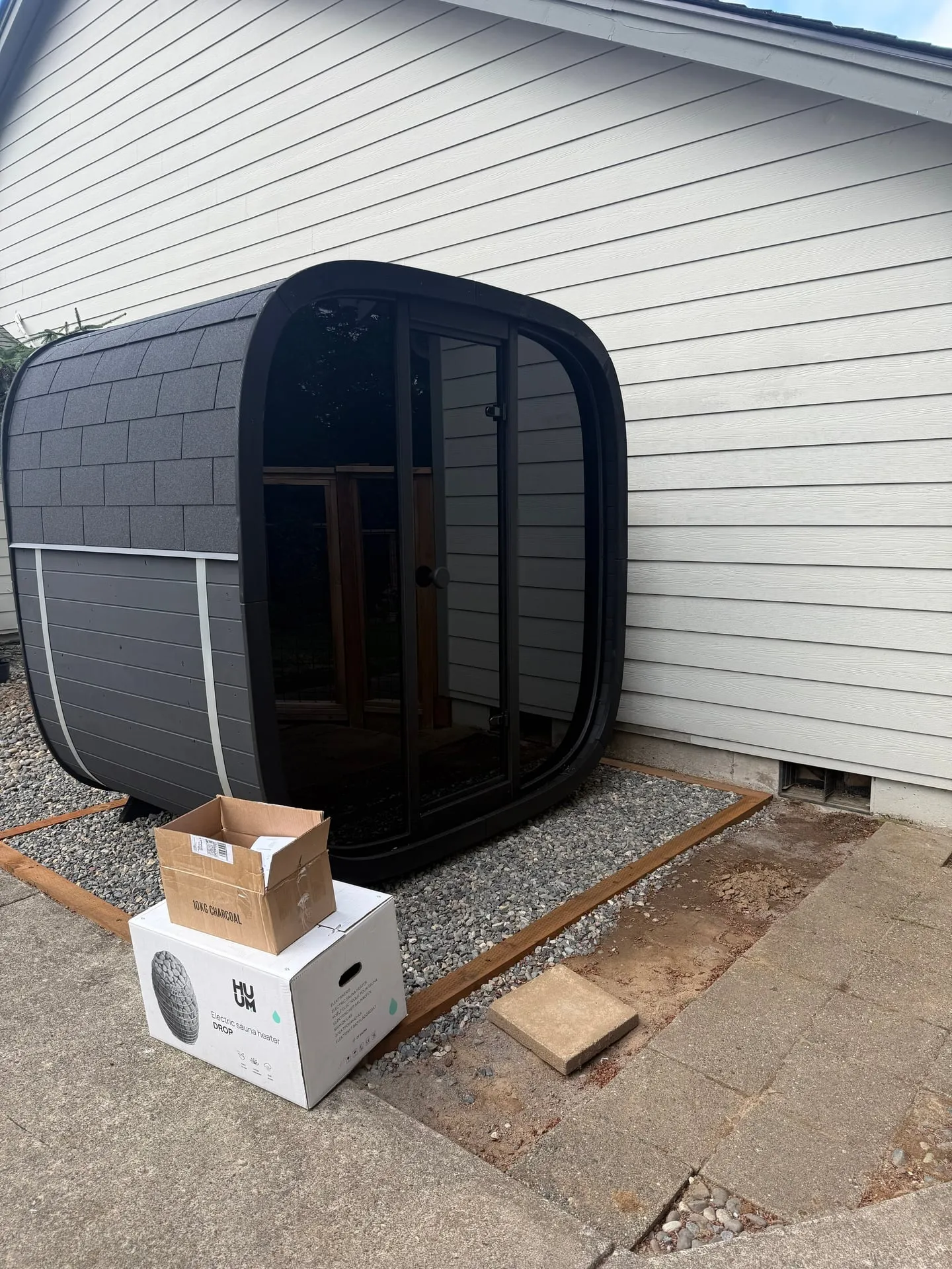 Freestanding black barrel-style outdoor sauna sitting on a gravel bed beside a white house in Washougal WA, with its cedar interior visible through the open door and the Huum heater box on the ground