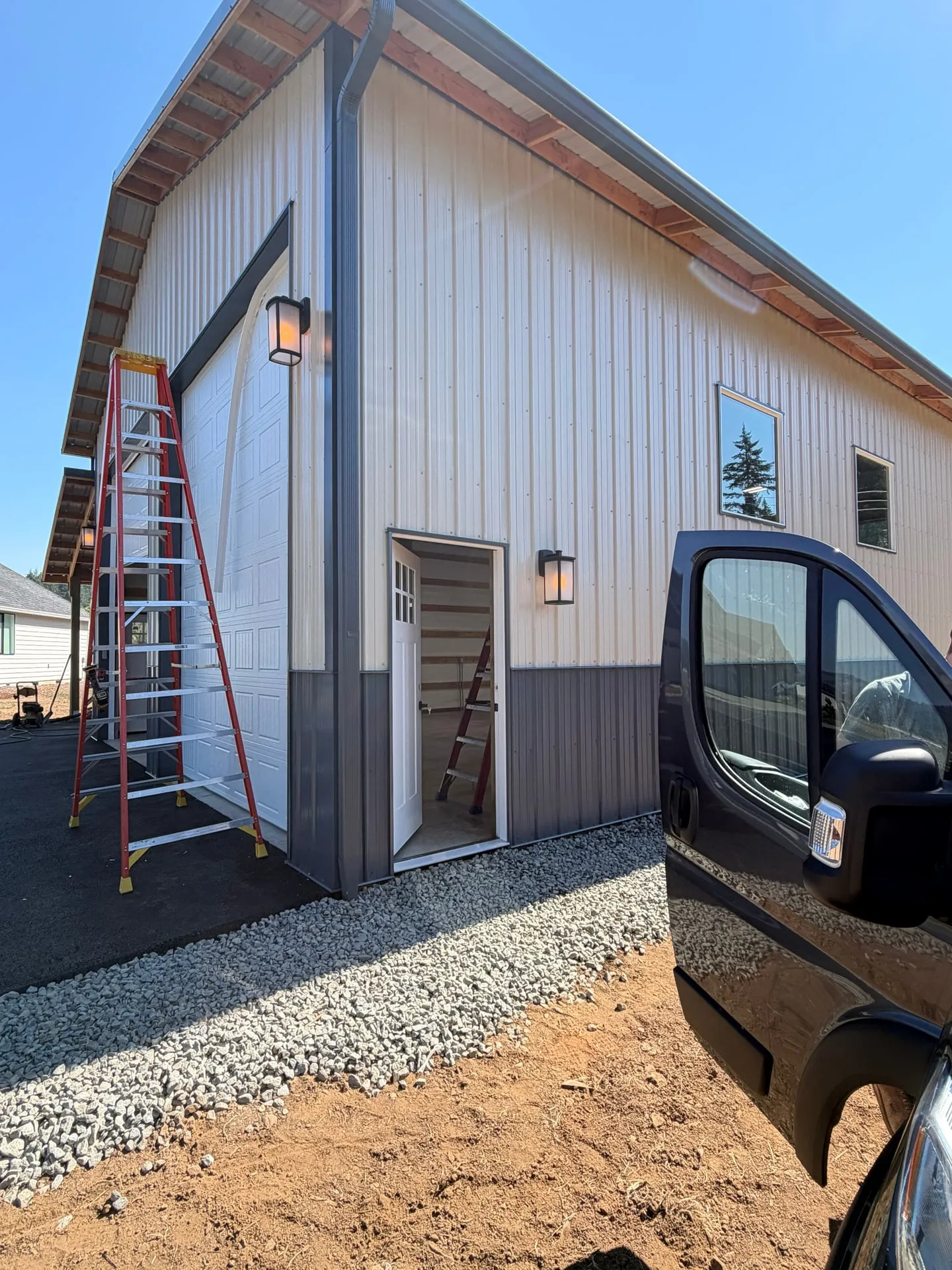 Exterior of a new white and grey pole barn in Battle Ground WA with a coach light mounted beside the personnel door and gravel apron in front of the overhead door