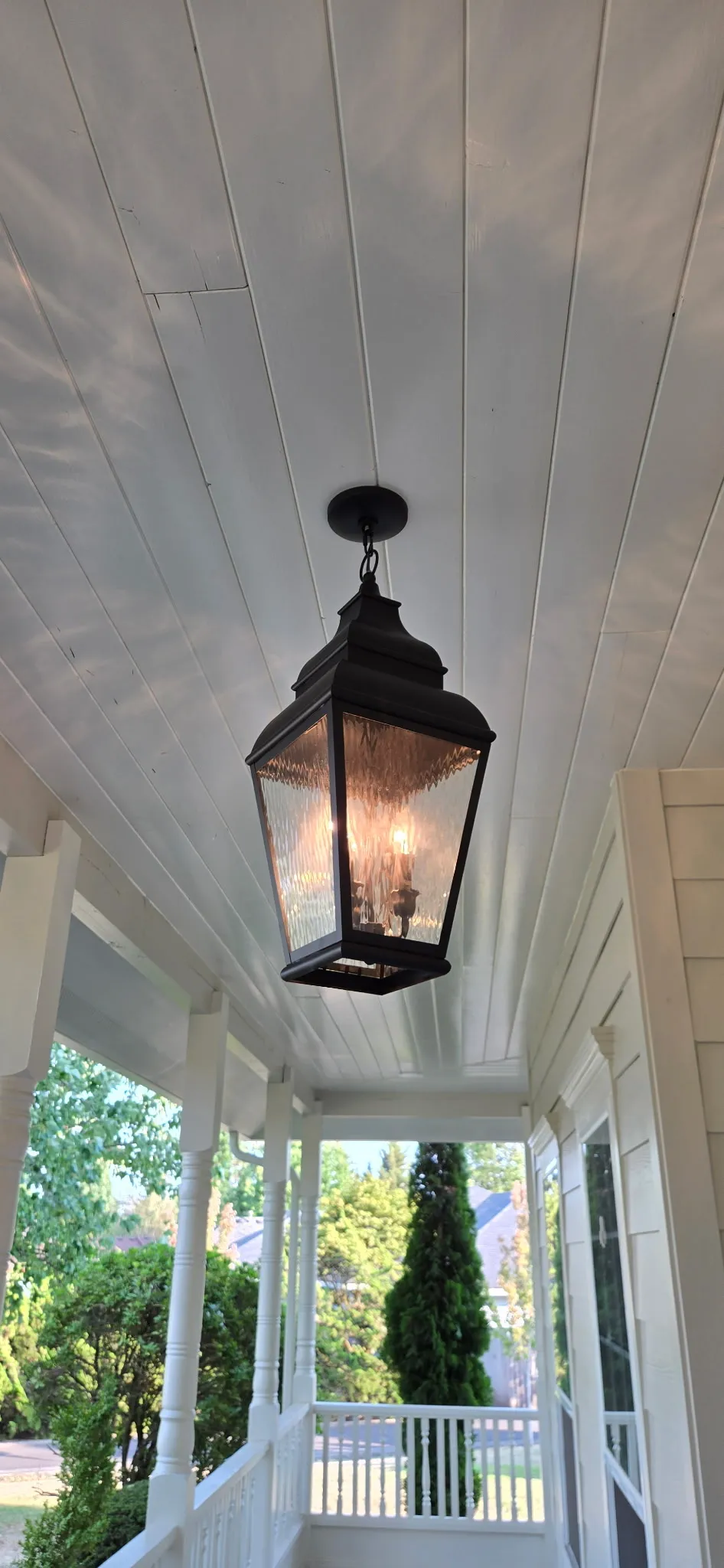 Black lantern pendant hanging from a pale blue shiplap porch ceiling on a custom home in Ridgefield WA, with white railings and the garden beyond