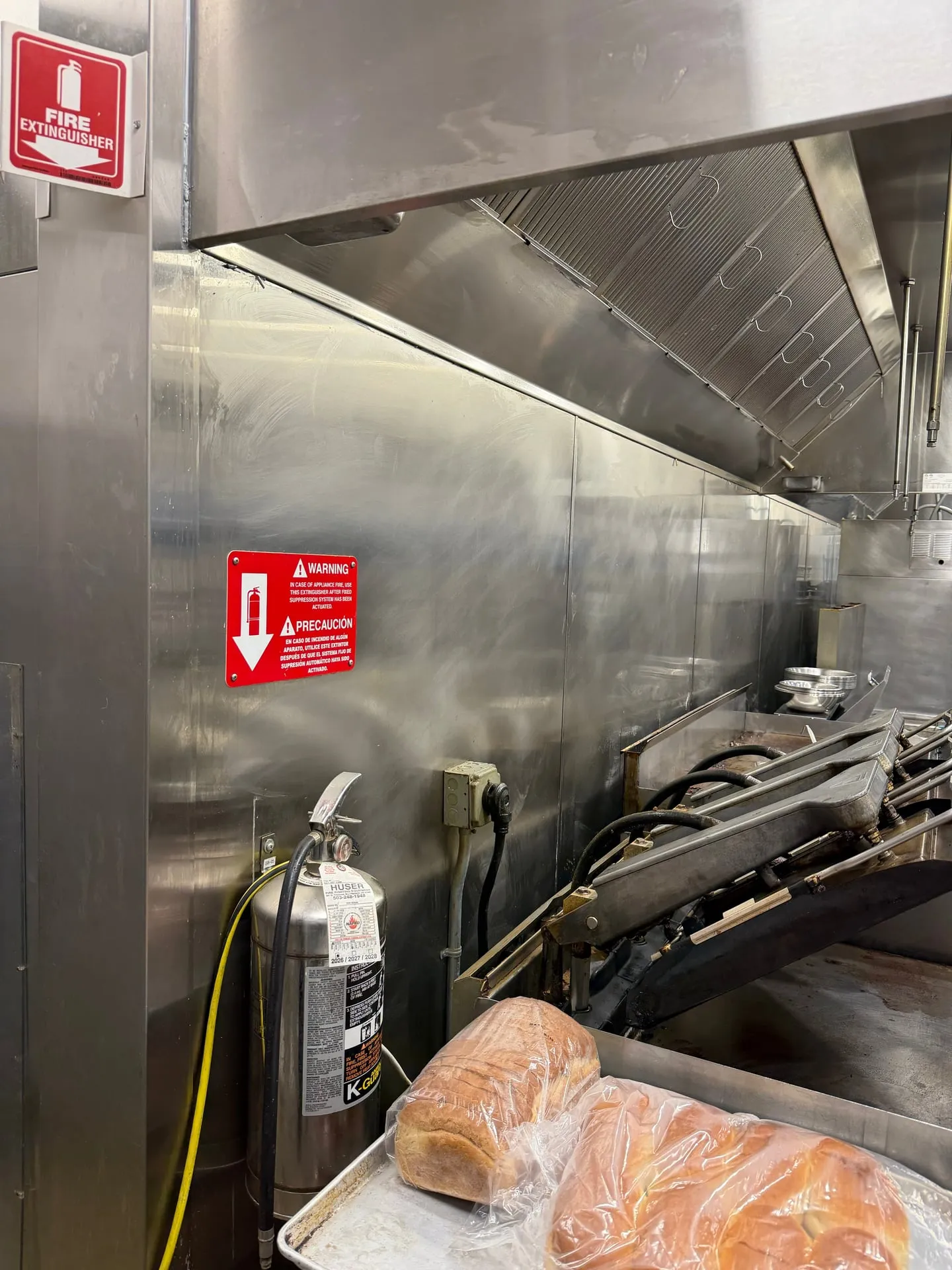 Restaurant cook line under a stainless steel hood in Ridgefield WA with a fire extinguisher, warning signs and an outlet on the back wall