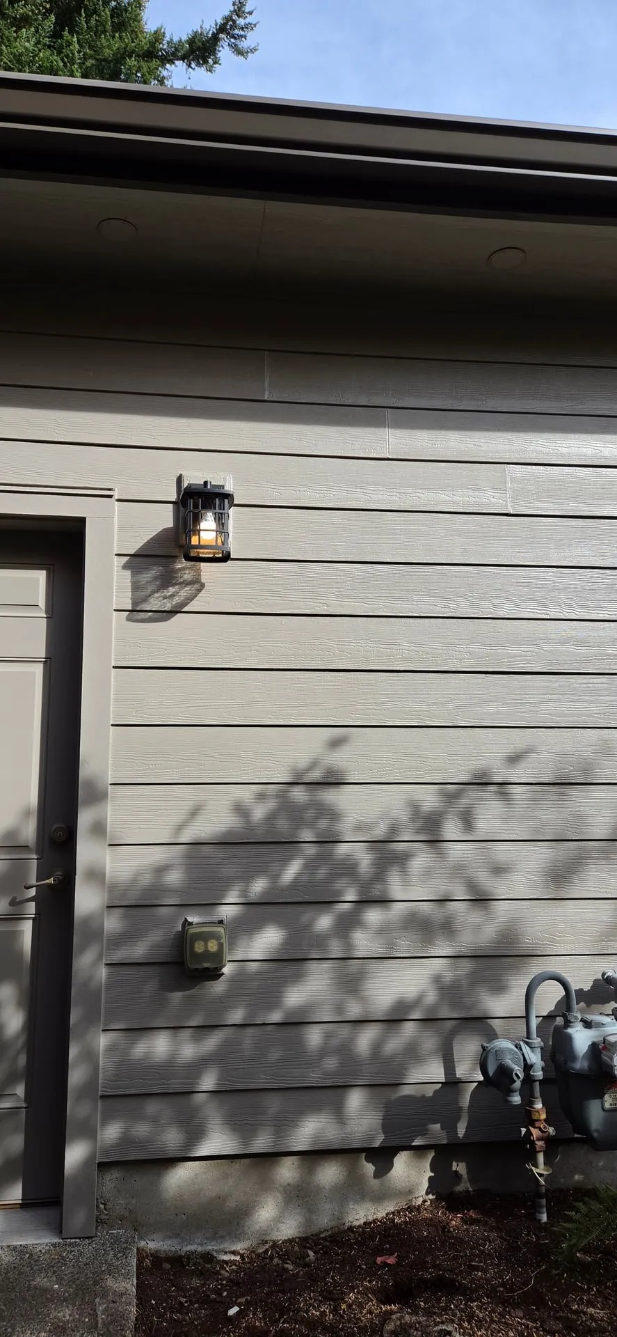 Black wall lantern lit beside a side door on gray lap siding of an east Vancouver WA home