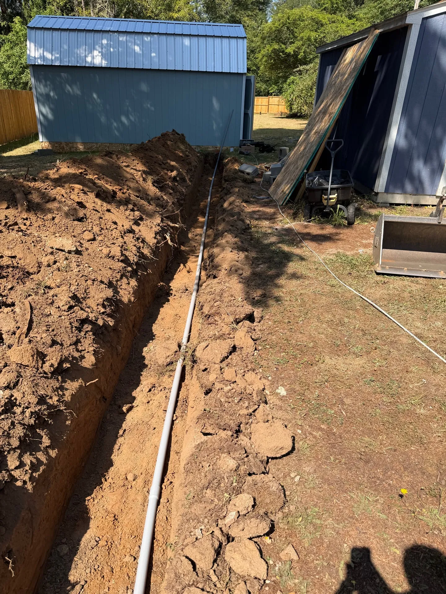 Open trench running across a rural Woodland WA backyard from the house toward a blue metal shed, with grey PVC conduit laid in the bottom of the trench
