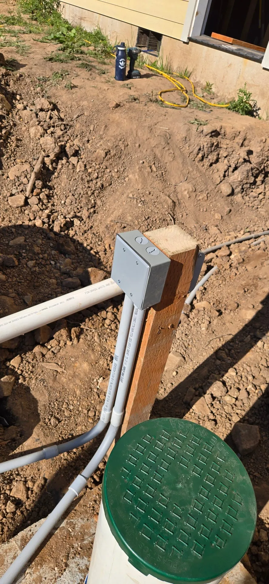 Grey underground conduit rising out of an open trench into a junction box on a post beside a green septic tank riser lid on a Kalama WA property
